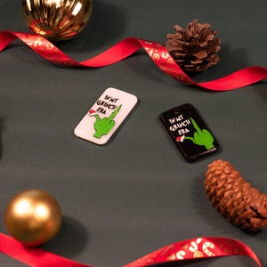 Puede incluir: Dos adornos navideños con una mano verde del Grinch y el texto "In My Grinch Era". Un adorno es blanco con texto rojo y el otro es negro con texto blanco.
