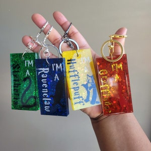 以下が含まれることがあります： グリッターが樹脂に埋め込まれたハリーポッターの家のキーホルダー4個。各キーホルダーには、家の紋章、動物のマスコット、白い文字で「私は[家の名前]です」というテキストが書かれています。キーホルダーは緑、青、黄、赤です。