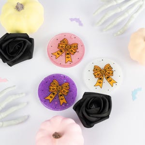 Può includere: Ciondoli in resina a tema Halloween con un design a fiocco e dettagli di ragni. I ciondoli sono rosa, viola e bianchi, con dettagli neri e arancioni. L'immagine include anche rose nere, zucche e una mano di scheletro.