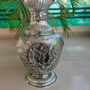 May include: Ornate silver vase with intricate floral and figure carvings. The vase features a wide base, a bulbous body, and a flared top. The detailed design includes a central relief of a person playing an instrument.