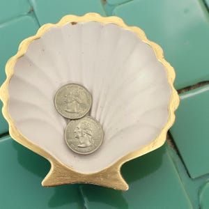May include: A seashell-shaped trinket dish with a gold-painted rim and base. The interior is a pale pink color, and it holds two silver quarter coins. The dish sits on a turquoise tile surface.