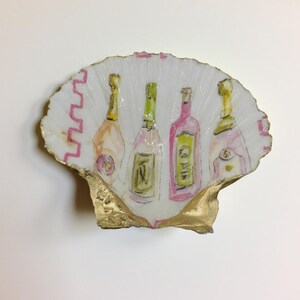 May include: A decorative seashell with a gold-painted edge, featuring a watercolor painting of four wine bottles in shades of pink, green, and gold. The shell has a white background with a pink zigzag pattern.