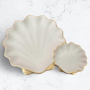 May include: A decorative, off-white seashell-shaped serving dish with a gold-colored trim. The dish has a large, scalloped shell shape connected to a smaller shell. The surface has a glossy finish.
