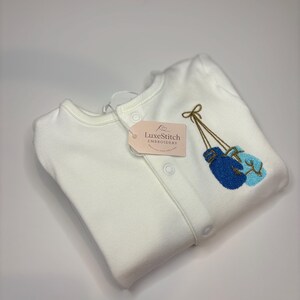 May include: A white baby sleepsuit with a button-up front and a tag that reads "LuxeStitch Embroidery." The sleepsuit features an embroidered design of two blue boxing gloves with gold-coloured strings.
