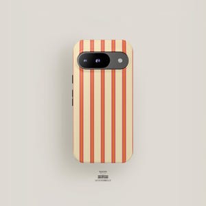 May include: A phone case with a cream-colored background and vertical orange stripes. The phone case has a camera cutout and side buttons. The text "MAISON ACCESSOBELLE" is at the bottom.