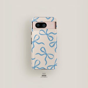May include: A cream-colored phone case with a blue bow pattern. The case has a raised camera bump with two lenses. The text "Maison Accessoirelle" is printed on the bottom of the case.