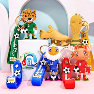 Könnte beinhalten: Eine Sammlung farbenfroher Schlüsselanhänger mit Fußballmotiven. Die Schlüsselanhänger umfassen Tierfiguren, eine goldene Trophäe und einen Fußballschuh. Jeder Schlüsselanhänger hat ein Band mit dem Text "FIFA WORLD CUP".