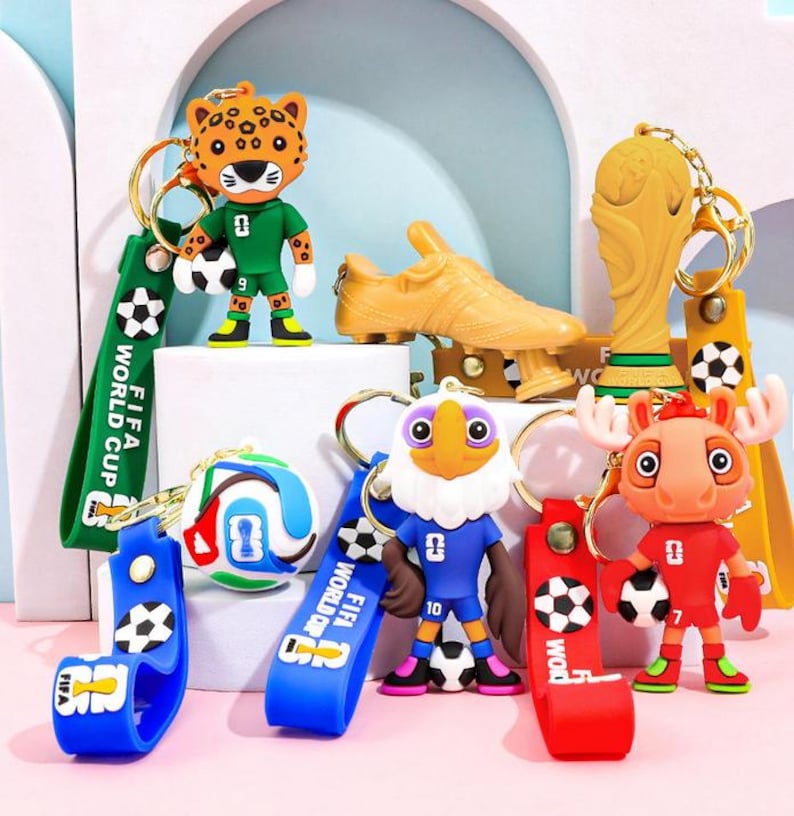 May include: A collection of colorful keychains featuring soccer-themed designs. The keychains include a leopard, eagle, moose, and soccer ball figures, along with a golden trophy and a soccer cleat. Each keychain has a strap with the text "FIFA WORLD CUP".