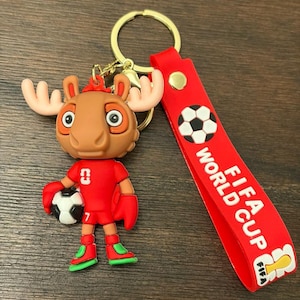 May include: A novelty keychain featuring a cartoon moose character in a red soccer uniform, holding a soccer ball. The keychain includes a red strap with "FIFA WORLD CUP" text and a soccer ball graphic. The moose has large antlers and is wearing green and red shoes.