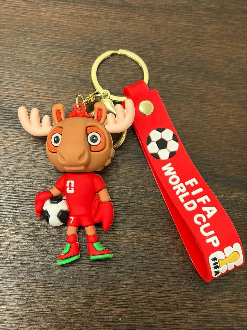 May include: A novelty keychain featuring a cartoon moose character in a red soccer uniform, holding a soccer ball. The keychain includes a red strap with "FIFA WORLD CUP" text and a soccer ball graphic. The moose has large antlers and is wearing green and red shoes.