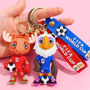 May include: Two novelty keychains featuring cartoon characters in soccer uniforms. One is a moose in a red uniform, the other an eagle in blue. Both hold soccer balls. Blue and red straps with "FIFA WORLD CUP" text are attached.
