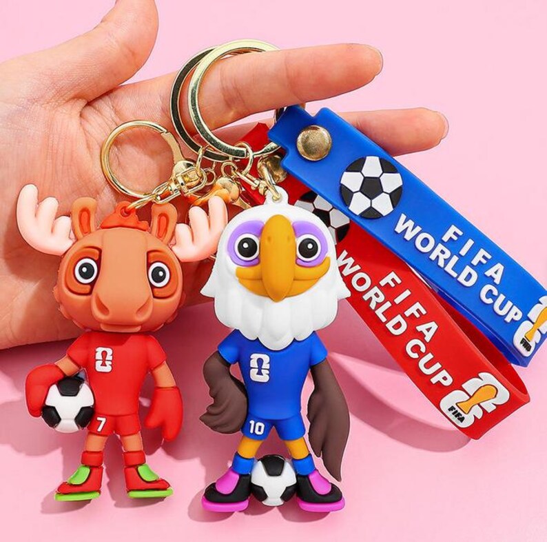 May include: Two novelty keychains featuring cartoon characters in soccer uniforms. One is a moose in a red uniform, the other an eagle in blue. Both hold soccer balls. Blue and red straps with "FIFA WORLD CUP" text are attached.