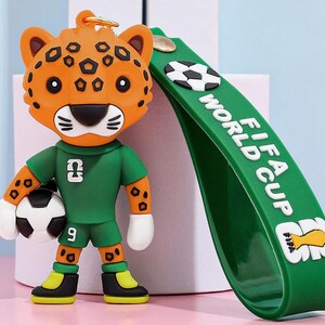 May include: A keychain featuring a cartoon jaguar wearing a green soccer uniform and holding a soccer ball. The jaguar has orange fur with black spots. A green strap with "FIFA WORLD CUP" text and soccer ball graphics is attached.