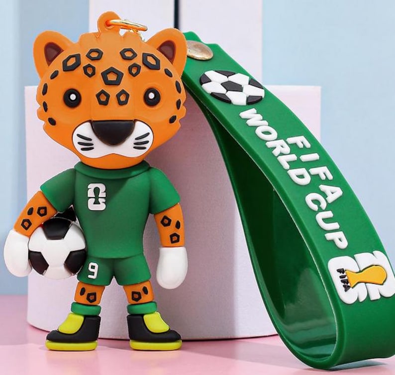 May include: A keychain featuring a cartoon jaguar wearing a green soccer uniform and holding a soccer ball. The jaguar has orange fur with black spots. A green strap with "FIFA WORLD CUP" text and soccer ball graphics is attached.