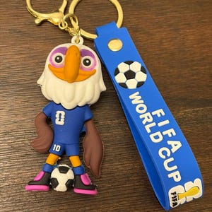 May include: A keychain featuring a cartoon eagle mascot in a blue soccer uniform, holding a soccer ball. The keychain has a blue strap with "FIFA WORLD CUP" text and a gold-colored clasp.