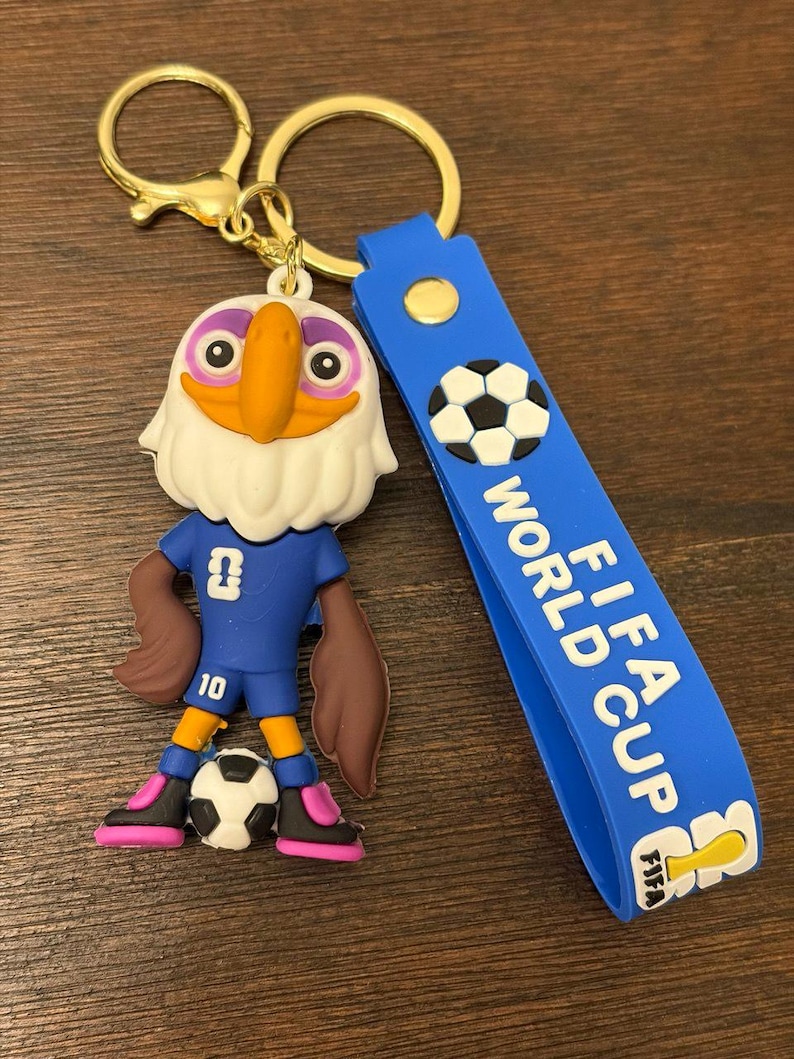 May include: A keychain featuring a cartoon eagle mascot in a blue soccer uniform, holding a soccer ball. The keychain has a blue strap with "FIFA WORLD CUP" text and a gold-colored clasp.