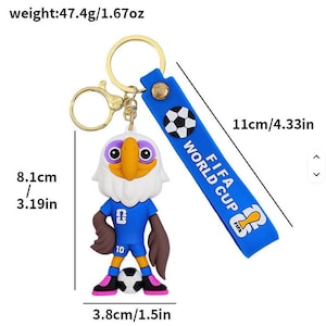 May include: A keychain featuring a cartoon eagle in a blue soccer uniform, holding a soccer ball. The keychain has a blue strap with "FIFA WORLD CUP" text and a soccer ball graphic. The eagle is 3.19 inches tall.