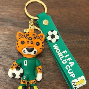 May include: A keychain featuring a cartoon jaguar mascot holding a soccer ball. The jaguar wears a green uniform with the number 9. The keychain has a green strap with the words "FIFA WORLD CUP" and a soccer ball graphic. The keychain is attached to a gold-colored ring.