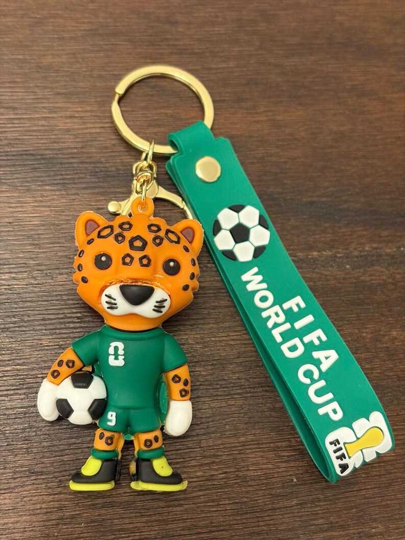 May include: A keychain featuring a cartoon jaguar mascot holding a soccer ball. The jaguar wears a green uniform with the number 9. The keychain has a green strap with the words "FIFA WORLD CUP" and a soccer ball graphic. The keychain is attached to a gold-colored ring.