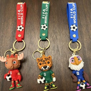 May include: Three keychains with the text "FIFA WORLD CUP" on the straps. Each keychain features a different cartoon mascot holding a soccer ball. The mascots are a moose, a jaguar, and an eagle. The straps are red, green, and blue.