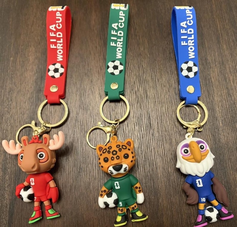 May include: Three keychains with the text "FIFA WORLD CUP" on the straps. Each keychain features a different cartoon mascot holding a soccer ball. The mascots are a moose, a jaguar, and an eagle. The straps are red, green, and blue.