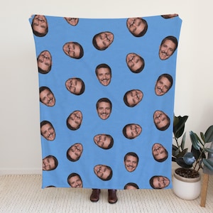 May include: A light blue blanket featuring a repeating pattern of smiling faces. The faces have brown hair and mustaches. The blanket is displayed in a room with a white door, a gray chair, and a potted plant.
