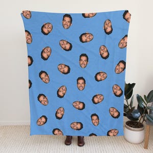 May include: A light blue blanket featuring a repeating pattern of a person's face. The faces are evenly spaced across the blanket. The blanket is made of a soft material and is suitable for use as a throw or bed covering.