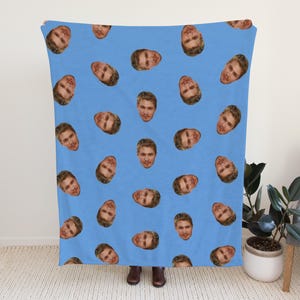 May include: A light blue blanket featuring a repeating pattern of a person's face. The faces are evenly spaced across the blanket. The blanket is held up in front of a person, with a neutral background.