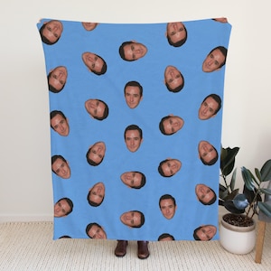 May include: A light blue blanket featuring a repeating pattern of a person's face. The faces are evenly spaced across the blanket. The blanket is held up in front of a person.