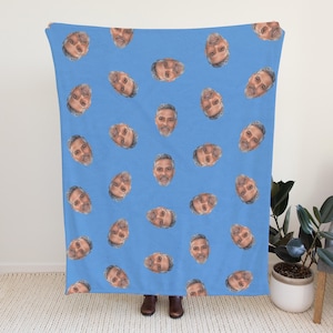 Könnte beinhalten: Eine hellblaue Decke mit einem sich wiederholenden Muster von Männergesichtern. Die Gesichter sind gleichmäßig über die Decke verteilt und erzeugen ein einzigartiges Design. Die Decke wird von einer Person gehalten.