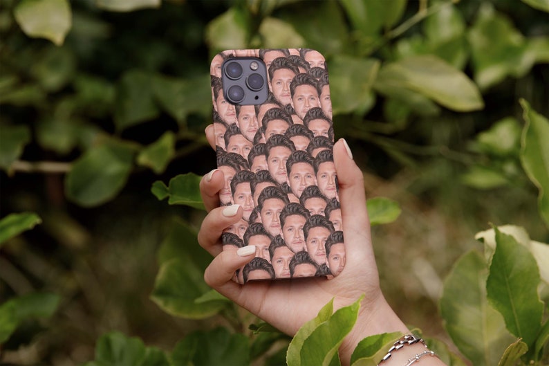 Puede incluir: Una funda de tel&eacute;fono con un patr&oacute;n repetido de la cara de una persona. La funda se sostiene en una mano sobre un fondo de hojas verdes. La funda tiene un acabado brillante y un recorte para la c&aacute;mara.