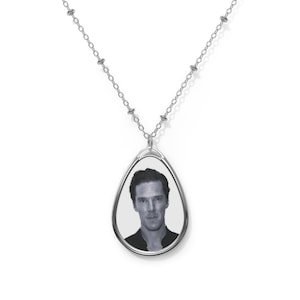 Puede incluir: Un collar plateado con un colgante en forma de lágrima. El colgante muestra un retrato en blanco y negro. La cadena es delicada, con pequeñas cuentas. El colgante está enmarcado en metal plateado.