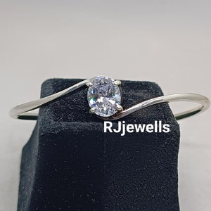 May include: A silver bangle bracelet with a single oval-shaped clear stone set in the center. The bracelet is on a black velvet display stand. The text "RJewells" is visible on the stand.