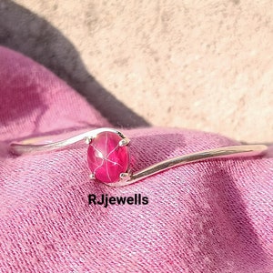 May include: A silver bangle bracelet with a pink ruby gemstone. The ruby is oval shaped and has a star-like pattern inside. The bracelet is curved and has a simple design.