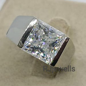 Peut inclure: Une bague en argent avec une grosse pierre précieuse claire de taille carrée. La bague a un design moderne avec un anneau épais et un sertissage à lunette. La pierre précieuse scintille, reflétant la lumière.