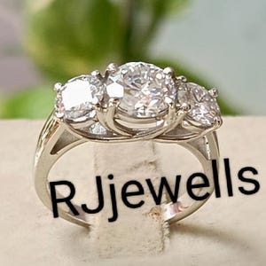 Könnte beinhalten: Ein silberner Ring mit drei klaren, runden Edelsteinen. Der Mittelstein ist größer als die beiden seitlichen Steine. Der Ring liegt auf einer neutralfarbenen Oberfläche, mit dem Text "Rjjewells" auf dem Ringband.