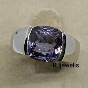 Puede incluir: Un anillo de plata con una gran piedra preciosa lavanda de talla cuadrada. La piedra preciosa está engastada en una banda sencilla y moderna. El anillo se muestra sobre una superficie de tono neutro, con el texto "RJewells".