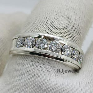 May include: A silver ring with a channel setting, featuring multiple round, clear gemstones. The ring is displayed on a white surface. The brand name "RJjewels" is visible on the ring.