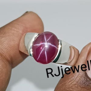 Może przedstawiać: Srebrny pierścionek z dużym, owalnym kamieniem szlachetnym w kształcie rubinu gwiazdy. Rubin ma głęboki czerwony kolor i wykazuje sześcioramienny wzór gwiazdy. Pierścionek ma prosty, nowoczesny design z szerokim pasmem.