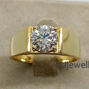 May include: A gold ring with a large, round, clear gemstone set in a rectangular bezel. The ring has a thick band and a polished finish. The gemstone sparkles brightly, reflecting light. The ring is displayed on a neutral-toned surface.