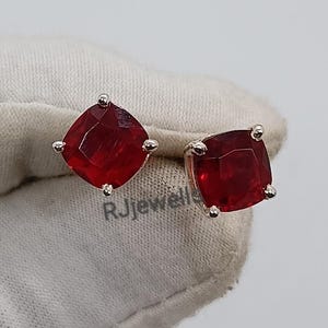 Puede incluir: Un par de pendientes de botón con piedras preciosas rojas cuadradas. Las piedras están engastadas en un metal plateado. Los pendientes se muestran sobre una superficie blanca. El texto "RJewells" es visible.