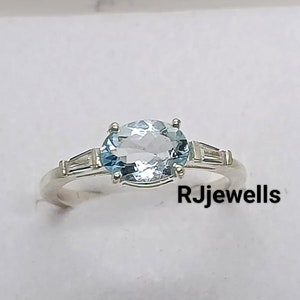 May include: A silver ring with a light blue oval aquamarine gemstone and two smaller baguette-shaped diamonds on either side. The ring is in a white box with the text "RJewells" on the bottom.