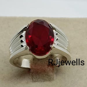 Könnte beinhalten: Ein silberner Ring mit einem großen, oval geschliffenen, tiefroten Edelstein. Der Ring hat ein klassisches Design mit einem gerippten Band auf beiden Seiten des Edelsteins. Der Ring wird auf einer neutralfarbenen Oberfläche präsentiert. Der Text "RjJewells" ist sichtbar.