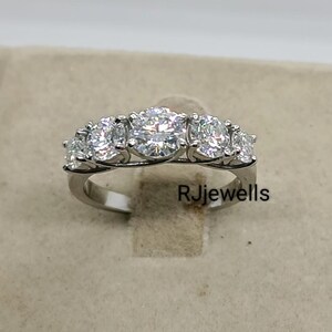 Peut inclure: Une bague en argent ornée de cinq pierres précieuses rondes et transparentes. La pierre centrale est plus grande que les autres. La bague est posée sur une surface de ton neutre. Le texte "RJjewells" est visible.
