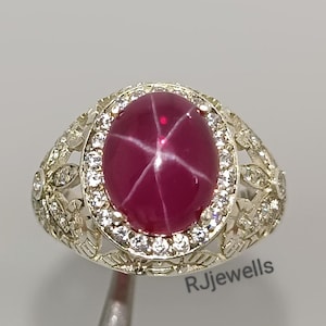 Puede incluir: Un anillo de plata con una gran gema de rubí estrella ovalada. El rubí está rodeado por un halo de pequeñas gemas transparentes. El anillo tiene un diseño calado y ornamentado en la banda. El texto "RJjewells" es visible.