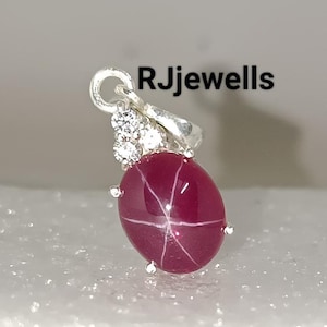 Könnte beinhalten: Ein silberner Anhänger mit einem roten Sternrubin-Edelstein mit weißen Akzenten. Der Anhänger ist auf einem weißen Hintergrund platziert.