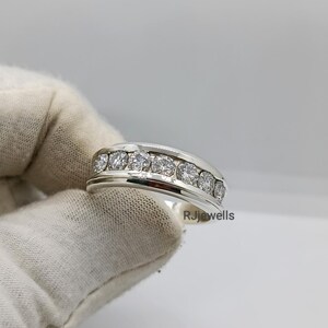 May include: A silver ring with a row of seven clear, round-cut stones. The ring has a polished finish and a wide band. The text "RJjewells" is visible on the side of the ring.