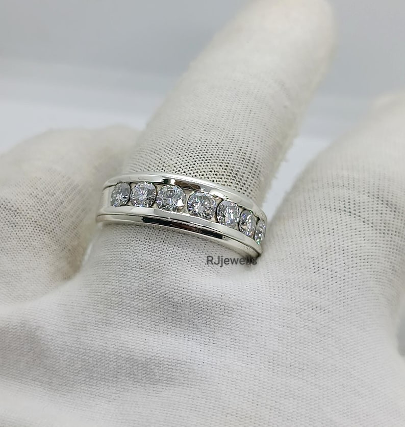 May include: A silver ring with a channel setting, featuring multiple round, clear gemstones. The ring is displayed on a white surface. The brand name "RJjewels" is visible on the ring.