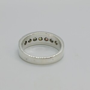 May include: A silver ring with a row of small, colorful gemstones. The ring is set against a plain white background, showcasing its polished metal and the vibrant hues of the stones.