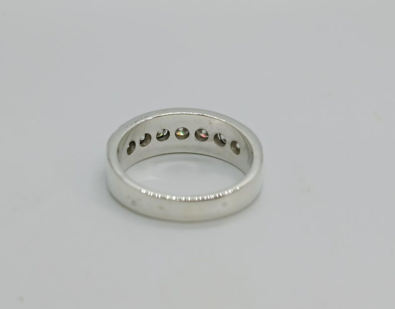 May include: A silver ring with a row of small, colorful gemstones. The ring is set against a plain white background, showcasing its polished metal and the vibrant hues of the stones.